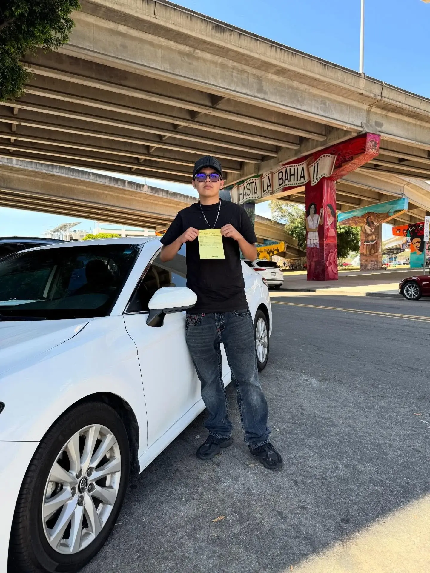 A Bonita Driving School student holding their certificate of completion beside the school car in Barrio Logan