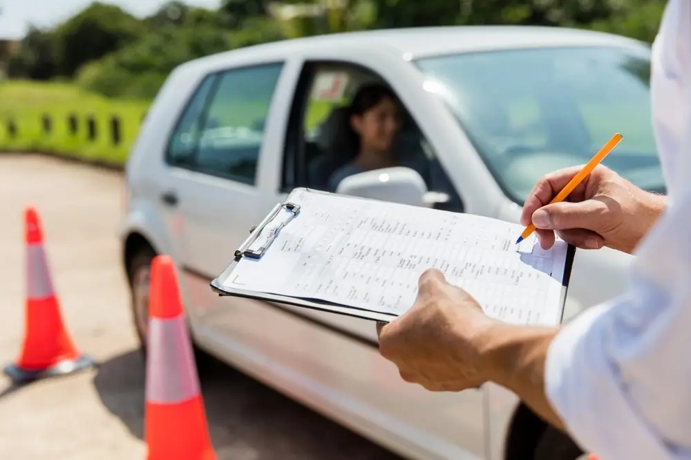 An examiner marking a drive-test score sheet beside a student waiting in the car