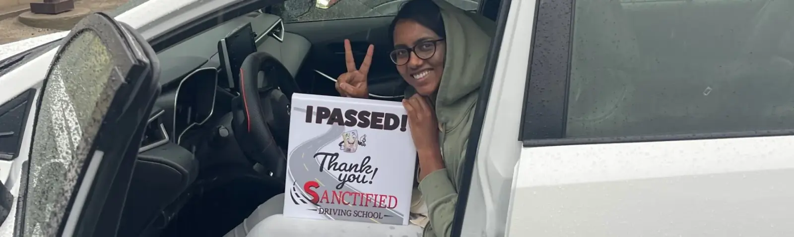 A Sanctified student in the driver’s seat holding an “I Passed!” sign